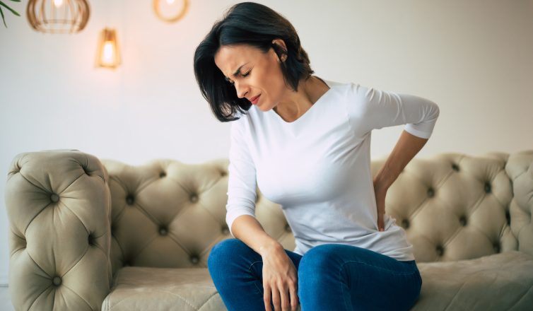 axial pain. close up photo of a hurting woman, who is sitting on a couch and holding her lower back with her left hand.