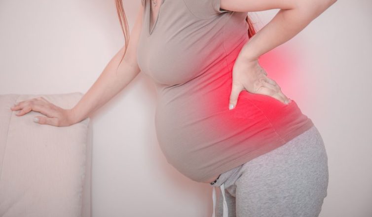 cropped view of young pregnant woman massaging her back. horizontal shape, side view, copy space. torso of pregnant woman with backache, pain in red.